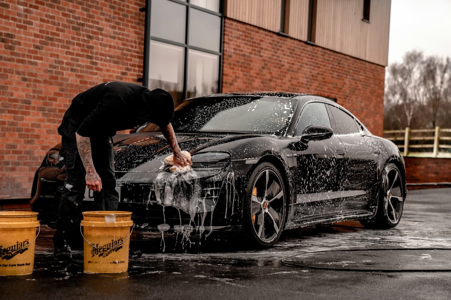 Petugas mencuci mobil hitam berbusa di halaman carwash, lengkap dengan ember dan selang